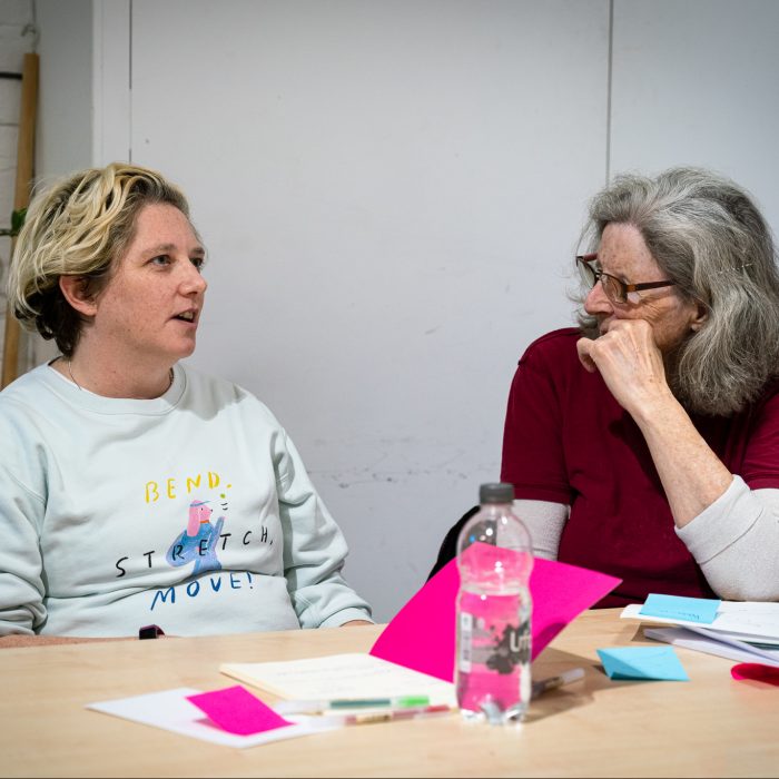 Two people sat talking in a workshop. 1 has short blonde hair and 1 has long grey hair and glasses.