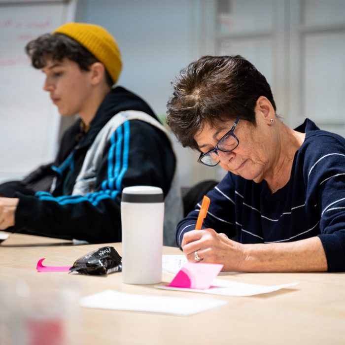 2 people sat writing in a writing workshop. 1 with short dark wavy hair under a yellow hat and 1 with short dark hair.