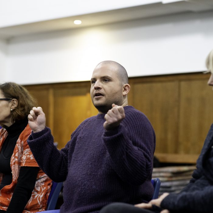 Three participants sat in a workshop. The one in the middle has a shaved head and stubble and is gesticulating whilst talking