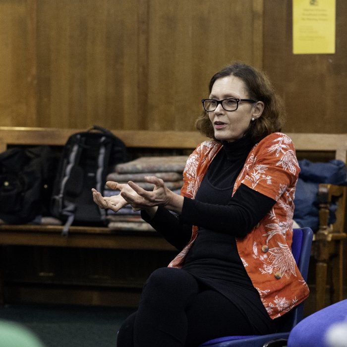 A person talking in a workshop. They have shoulder length brown hair and glasses and gesticulate whilst they talk.