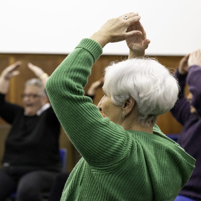 Five people doing a workshop activity. They are sat on chairs and all have their arms raised in a triangle above their heads.