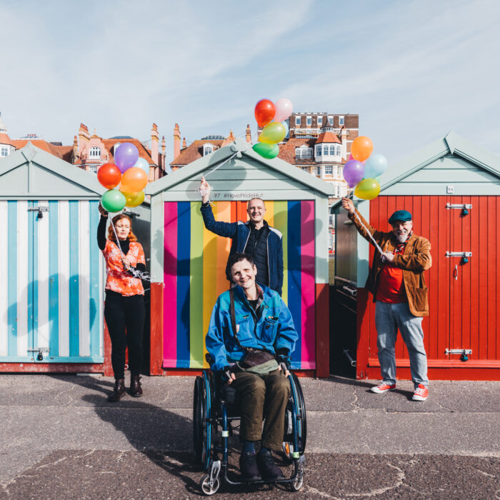 Four people stand in front of a rainbow coloured beach hut. They are holding colourful balloons and are smiling at the camera.